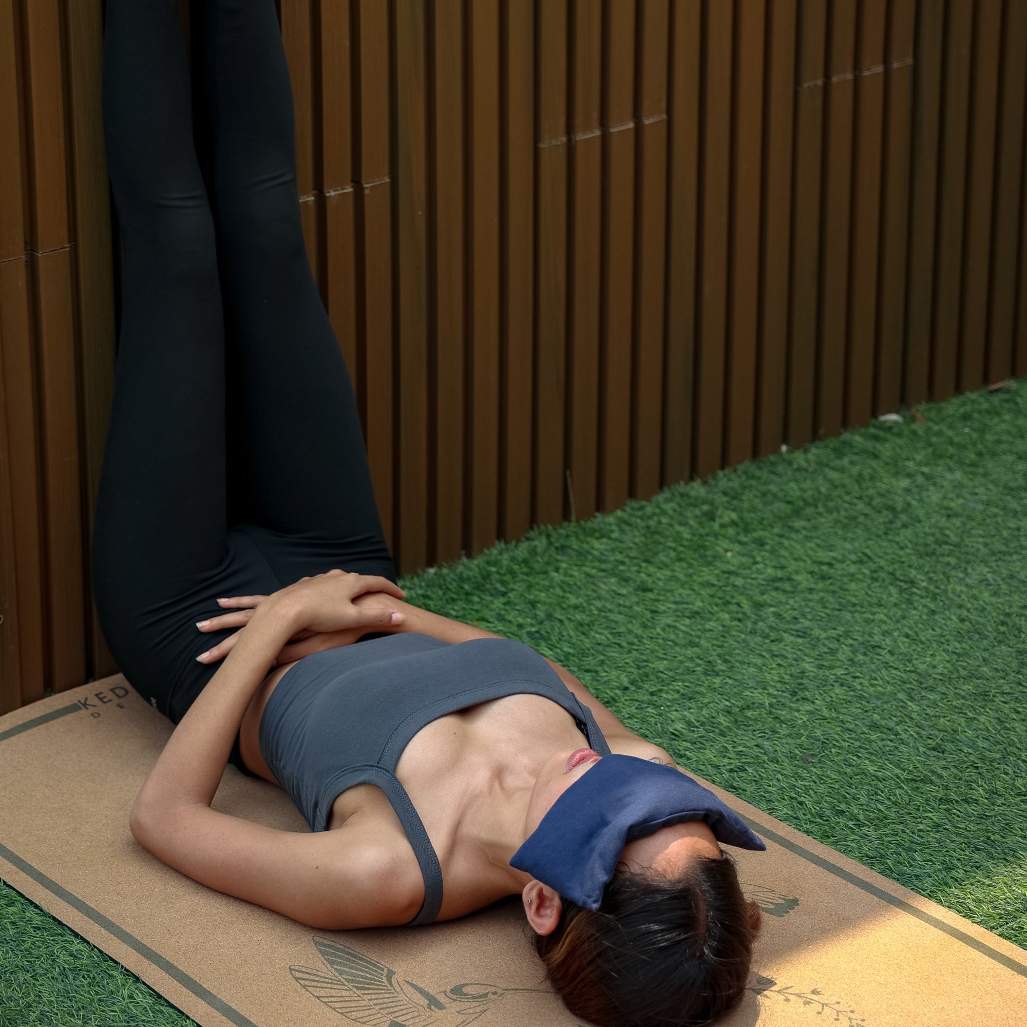 Person lying on a cork yoga mat wearing a kedgeree eye pillow against a wooden wall.