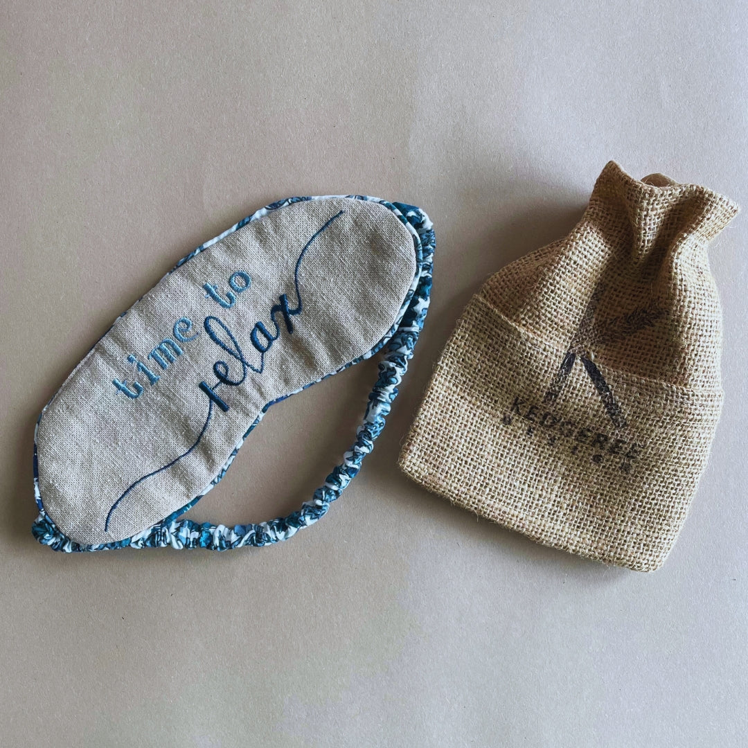 A gray and blue eye mask with the text 'Time to relax' on it, placed next to a burlap drawstring bag with the brand 'kedgeree' on it.