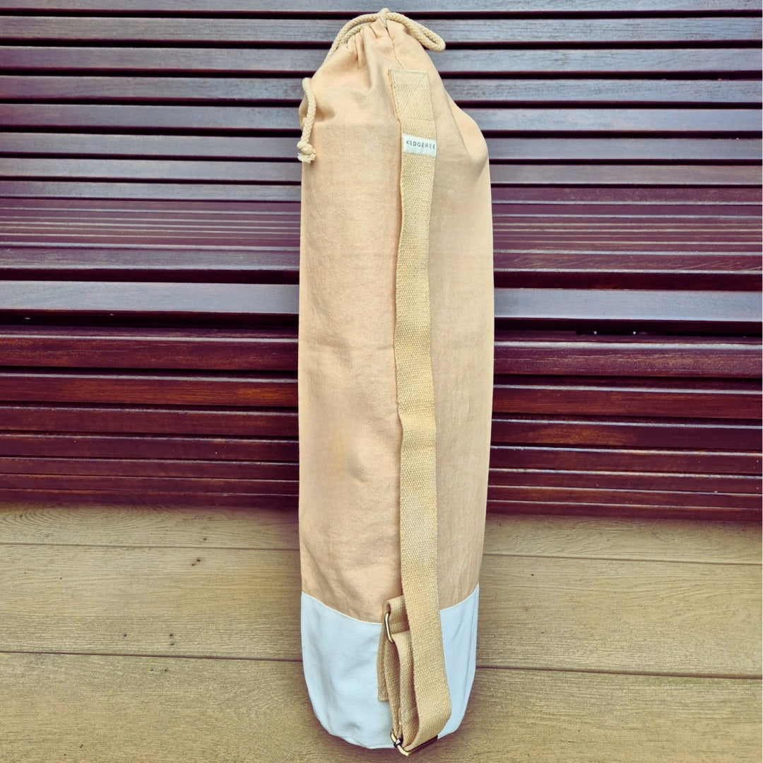 A beige travel yoga bag made of canvas cotton, standing upright on a wooden floor against a wooden background. It has a single shoulder strap and a drawstring top, with the Kedgeree Design logo visible.