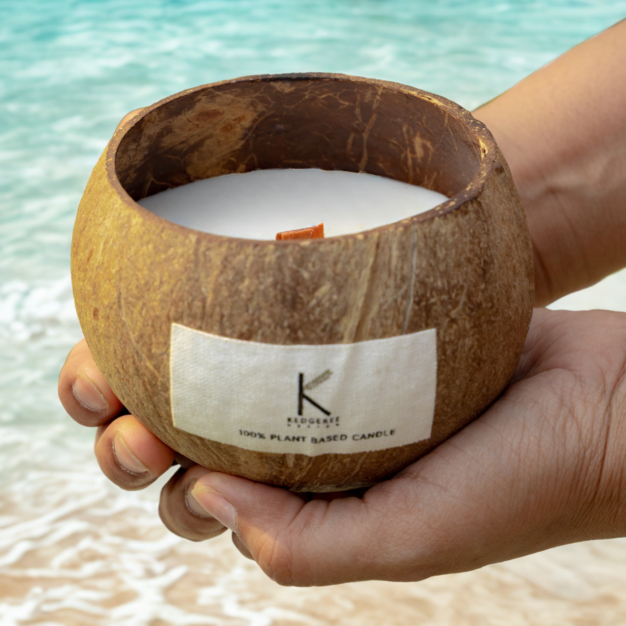 Vegan soy & coconut wax candle — virgin coconut and coconut milk. Plant Based Coconut shell candle with a label held by a hand against a beach background