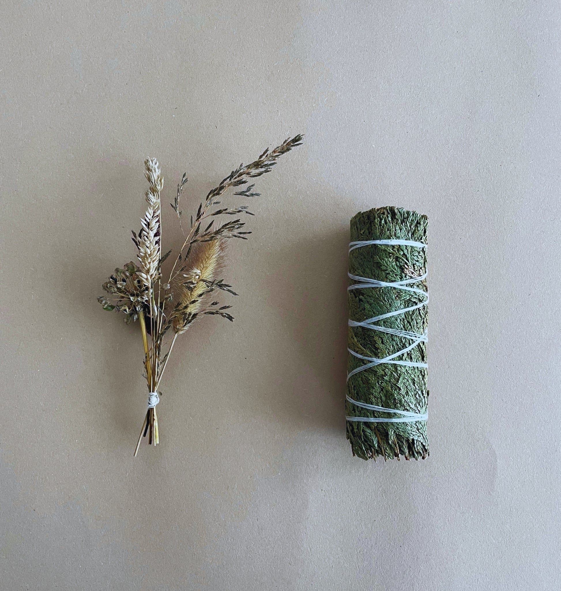 A handcrafted cedar smudge stick with tied dried flowers next to a handmade pouch, all placed on a beige background.