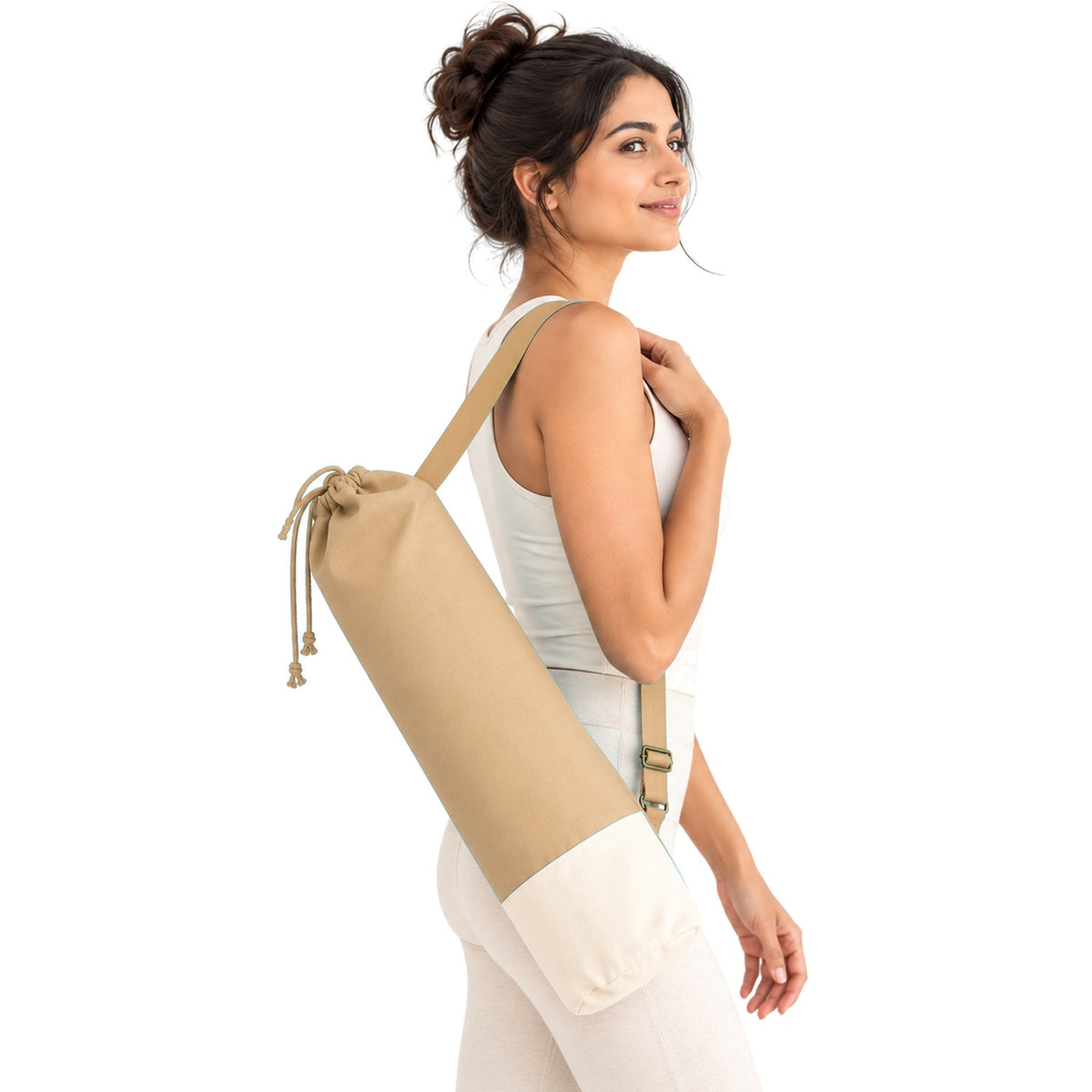 A woman in a white tank top and leggings carries a beige drawstring travel yoga mat bag over her shoulder against a plain white background.