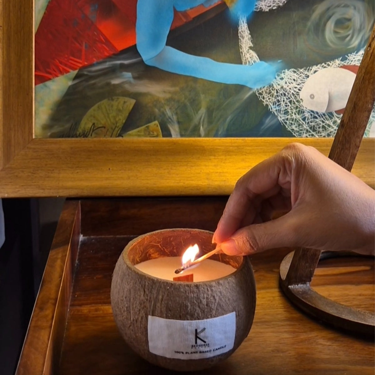 Coconut shell candle being lit with a match, placed on a wooden surface with a framed abstract painting in the background.
