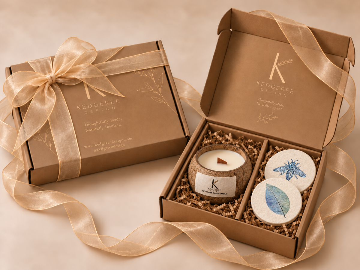 A gift set from Kedgeree Design featuring a brown kraft paper box tied with a beautiful ribbon, next to an open box containing a small coconut jar candle and two round decorative coasters and lid with blue and green leaf designs.