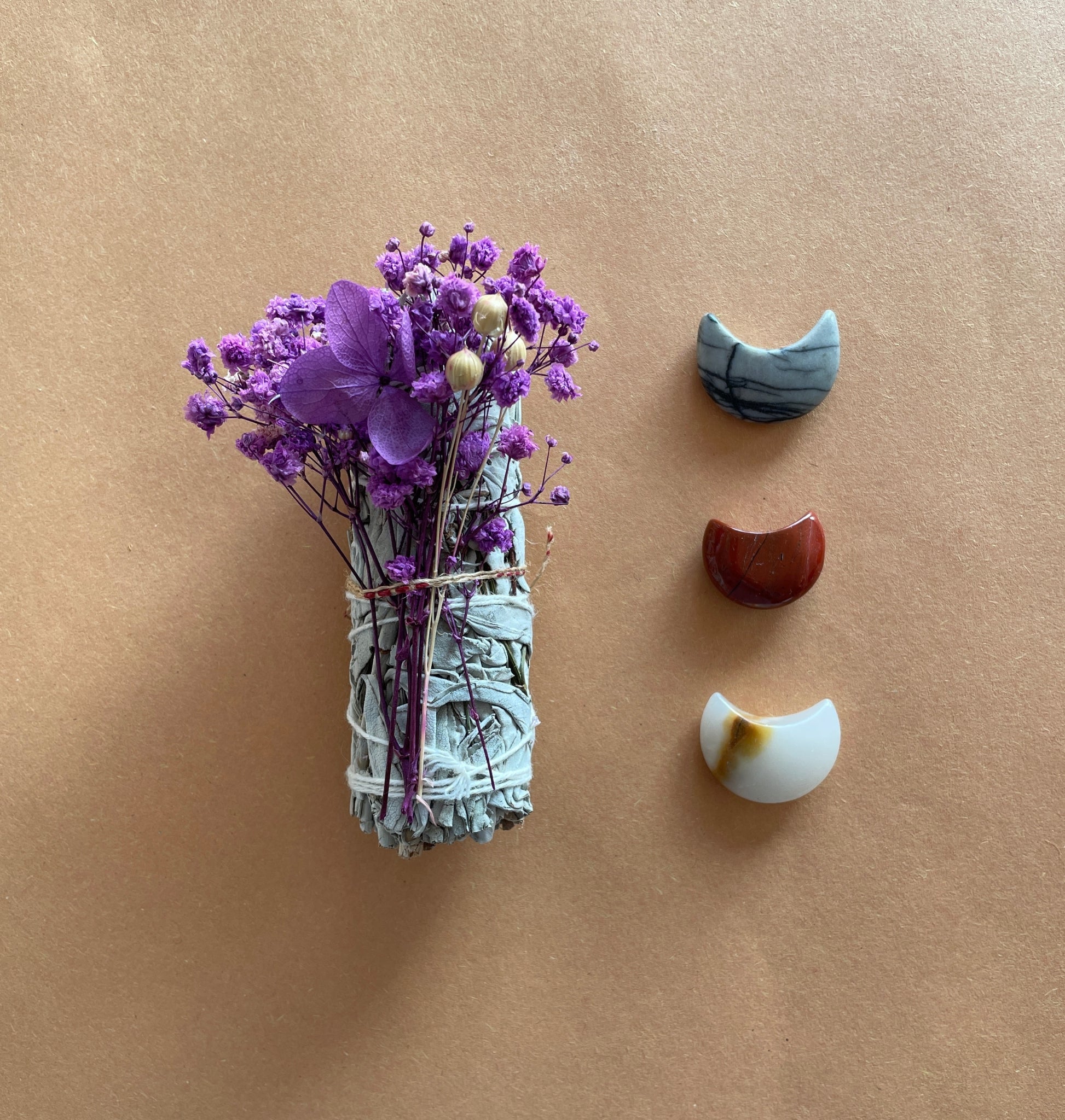 A bundle of white sage sticks wrapped in cotton string with purple flowers and decorative stones displayed on a brown surface.
