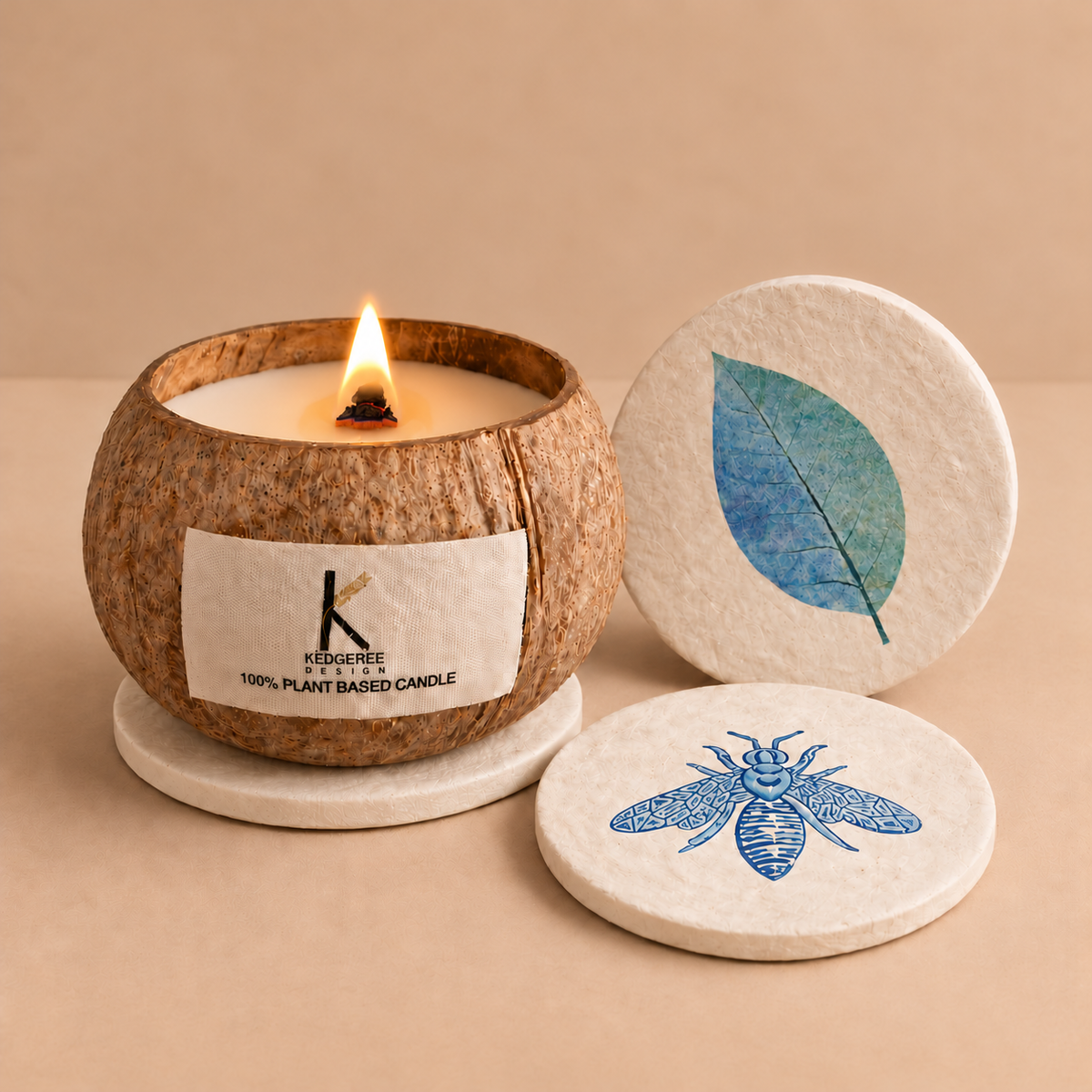 A lit KedgereeCandle in a natural coconut shell holder, shown with two round coasters and candle lid featuring a blue leaf and bee illustration on a warm beige background.