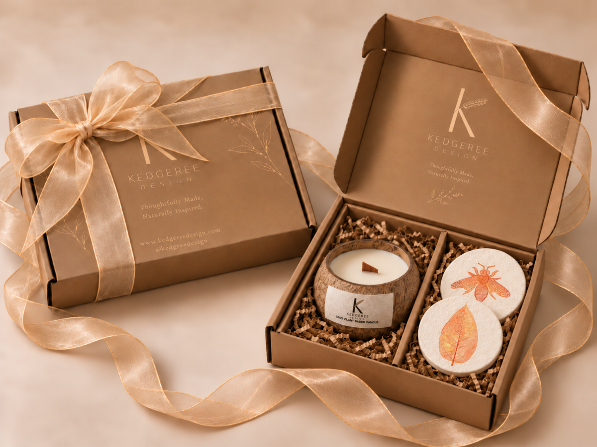 A Kedgeree gift set featuring a kraft paper box with a matching ribbon bow next to an open box containing a candle and two round decorative coasters and lid with botanical illustrations.