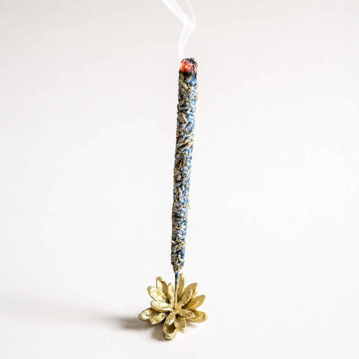 Kedgeree clean burn lavender incense sticks with blue and gold patterned wrapping, shown standing upright with a small white wick and visible smoke rising from the burning tip. The brass incense holder has a decorative gold-toned flower-like base on a clean white background.