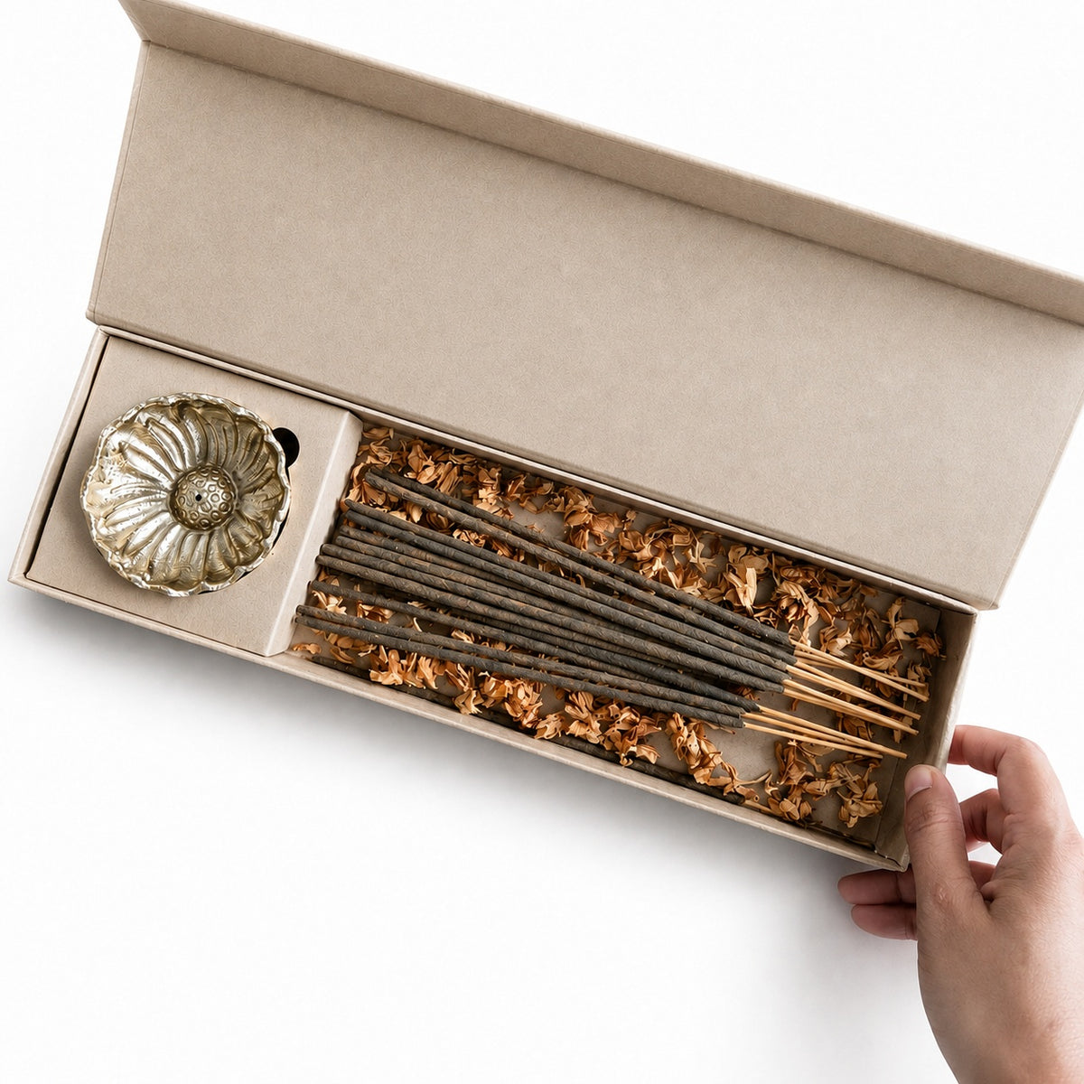 An open kraft paper gift box containing incense sticks nestled in dried flower petals and a small metallic incense holder, with a hand holding the box edge against a white background.
