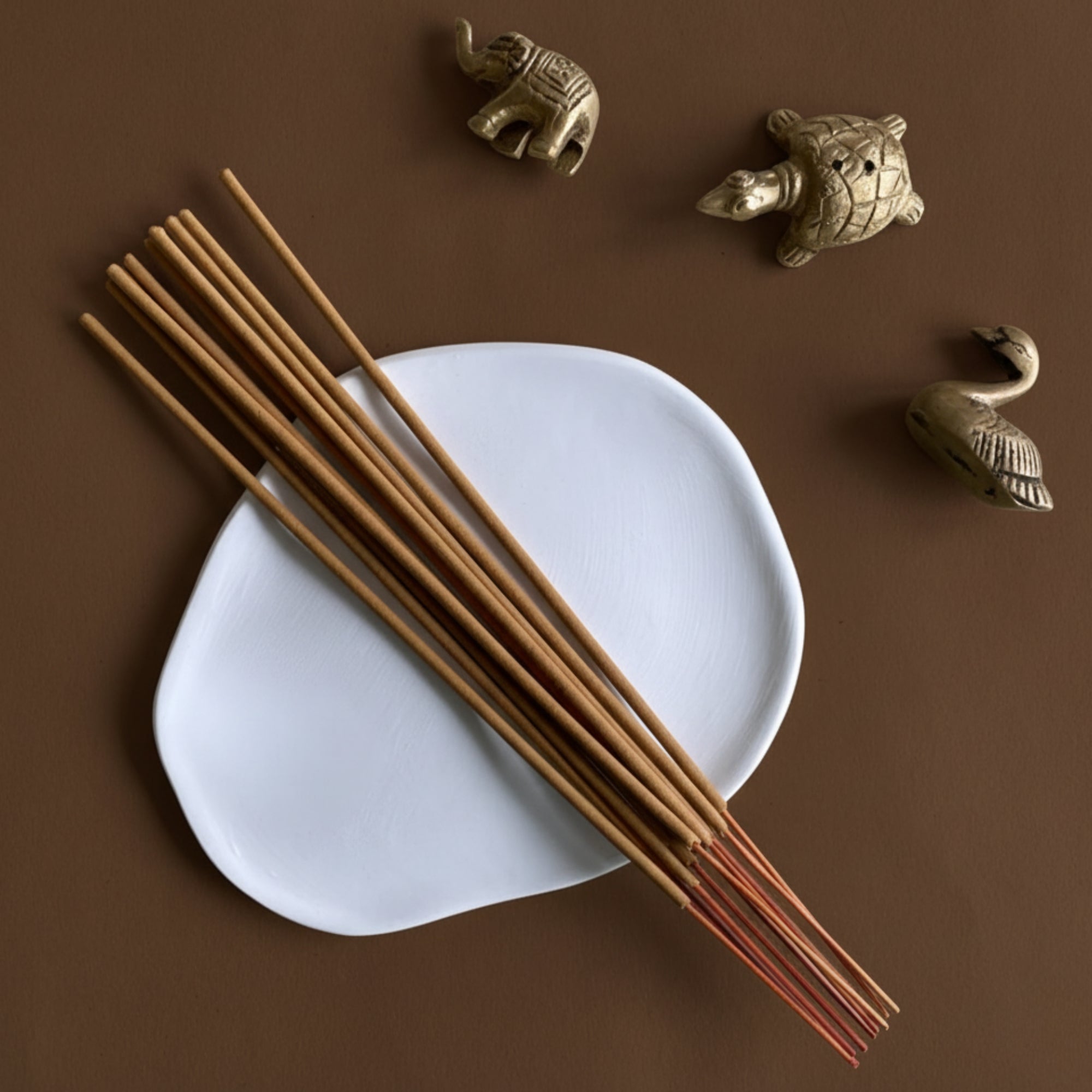 Sandalwood incense sticks on a white ash catcher tray with decorative handmade brass animal figurines incense holder on a brown background