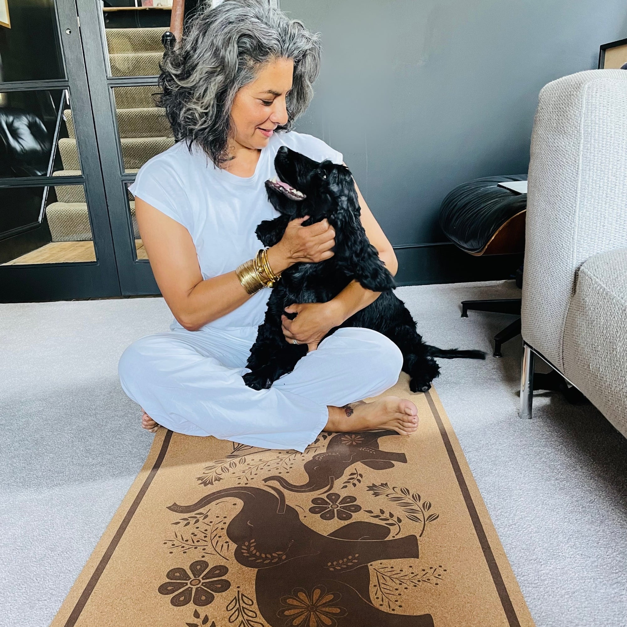 Woman holding a black dog on a yoga mat with a elephant design in a home setting. 