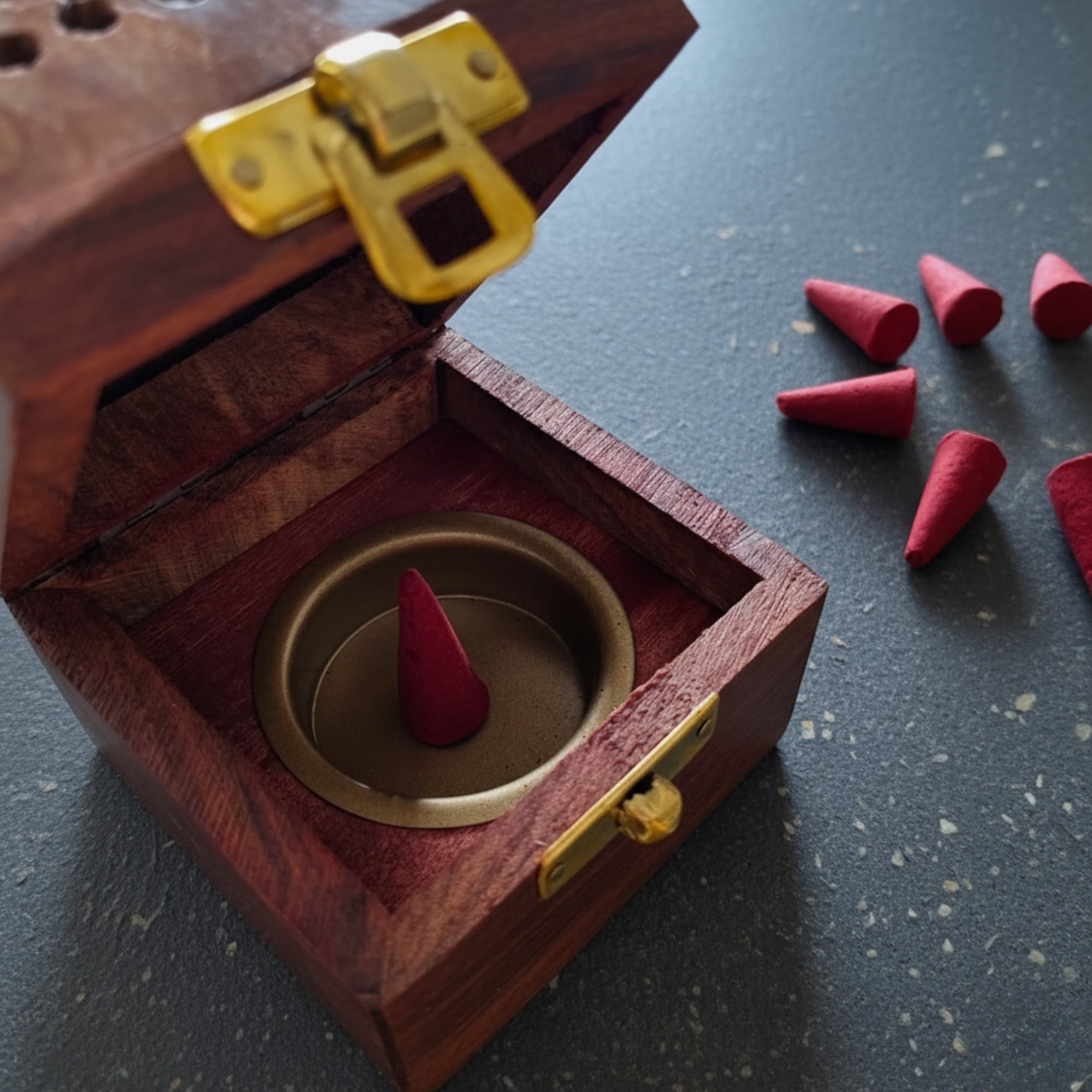 Wooden incense burner containing a red cone with cinnamon fragrance surrounded by more red cones on a dark surface.