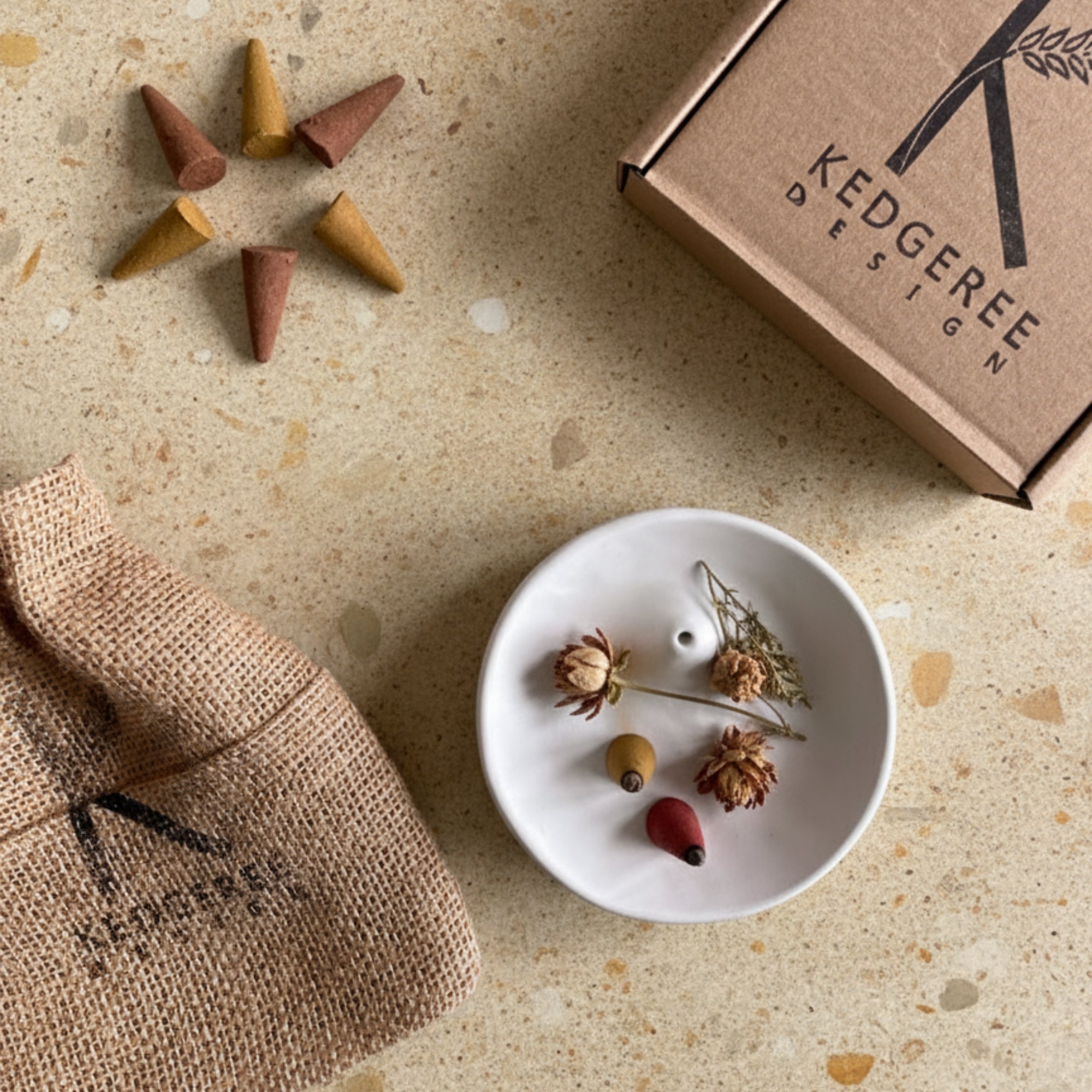 Incense cones, a white incense holder and palo santo burner, a brown gift bag, and a Kedgeree Design gift box on a textured surface.