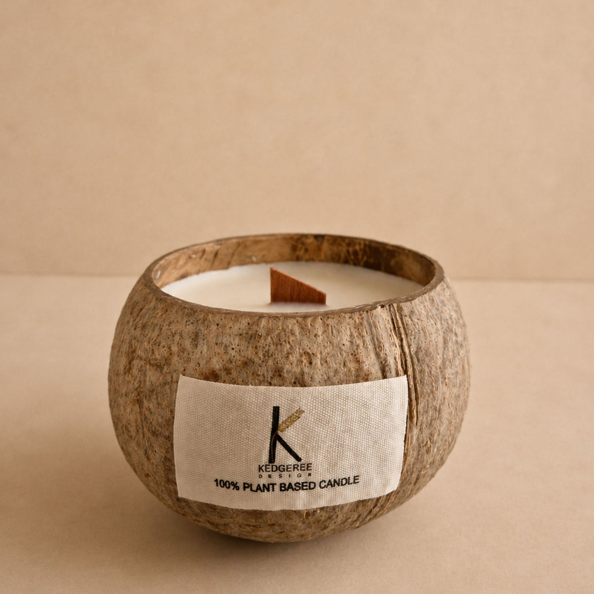 A coconut shell candle with a white wick and cream-colored wax, featuring a white fabric label with the brand name 'Kedgereese Design'. The candle is set against a neutral, solid beige background.