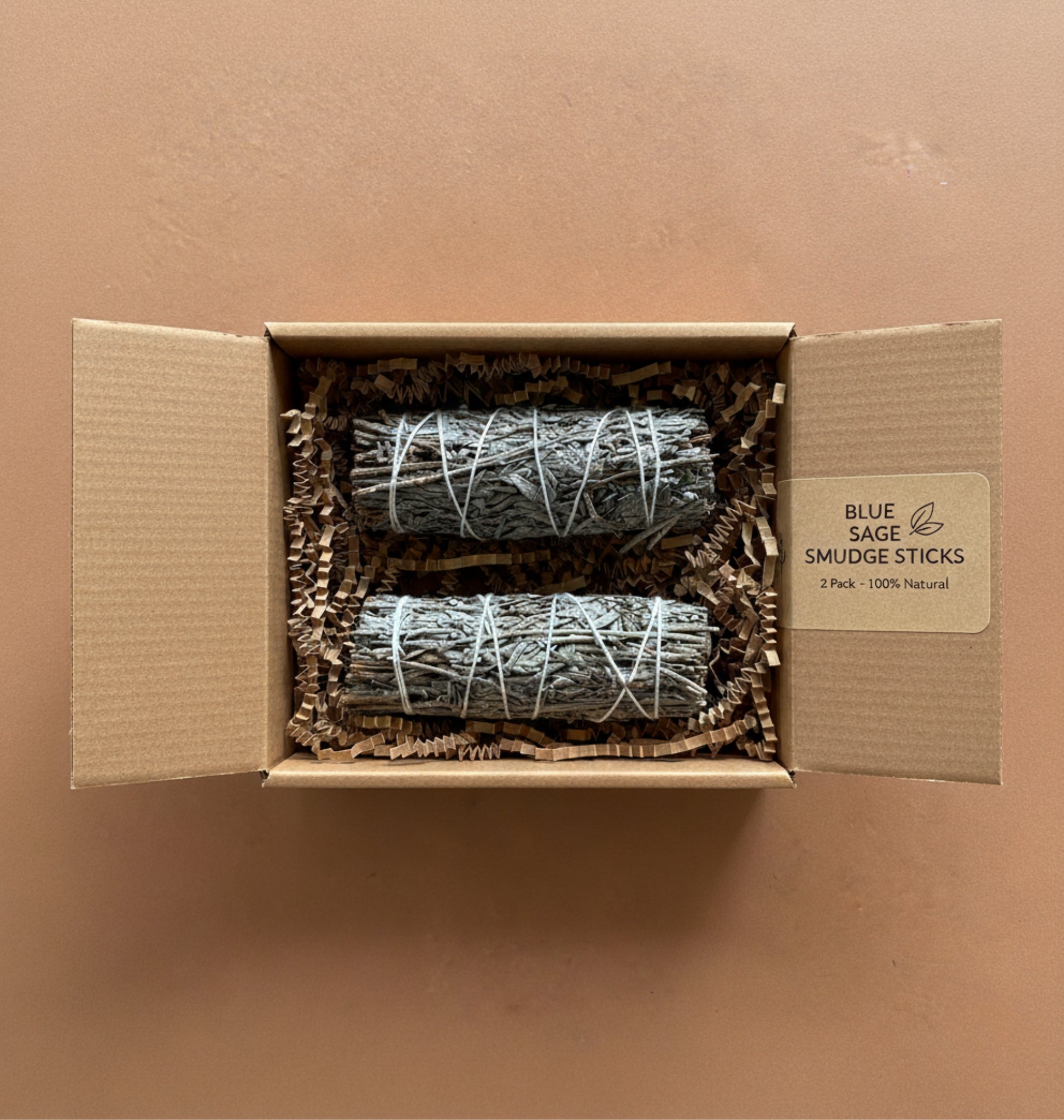 Two bundles of blue sage smudge sticks in a gift box with a label on a brown background.