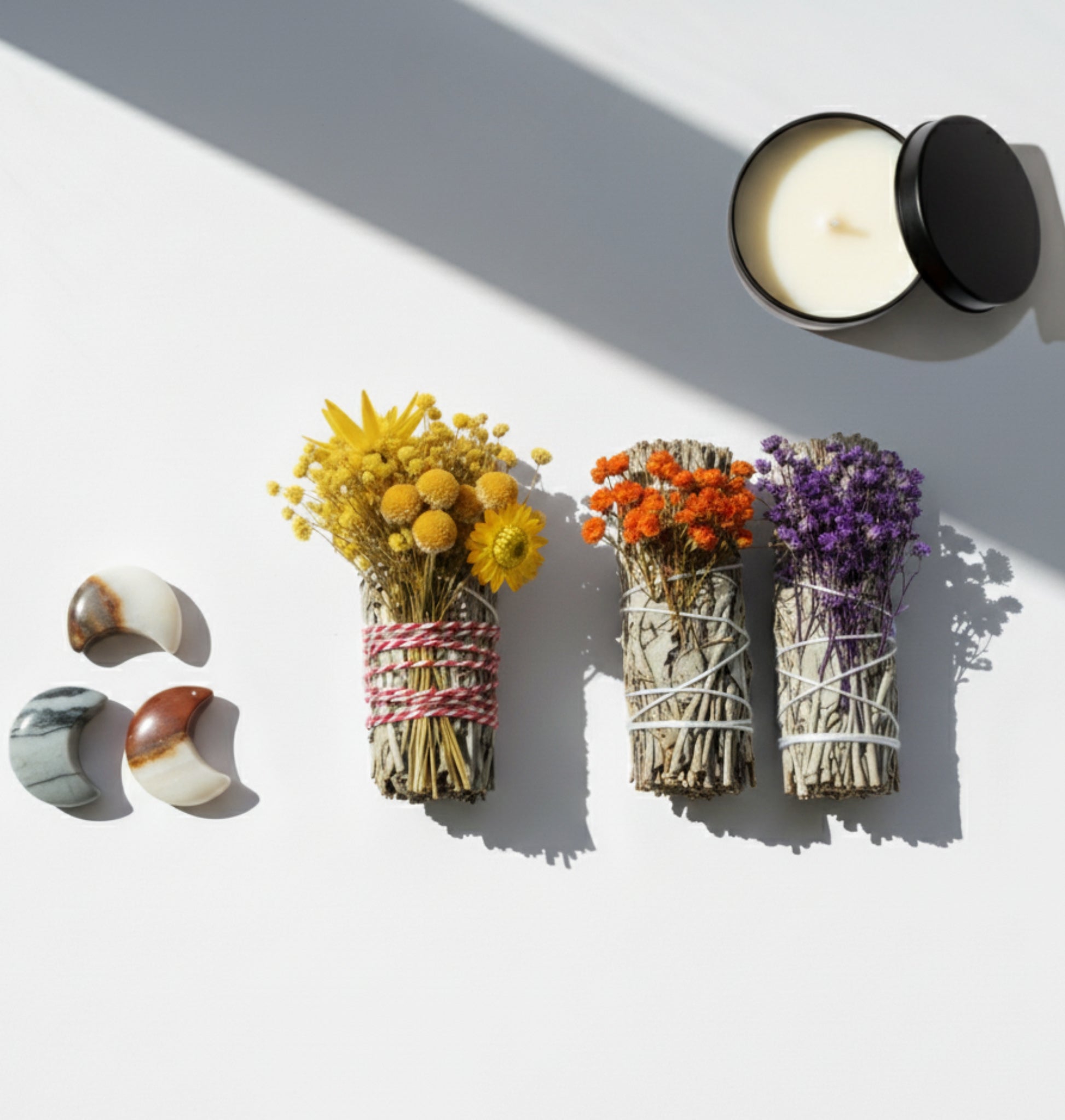 Three different natural smudge sticks with dried flowers and a candle on a white surface