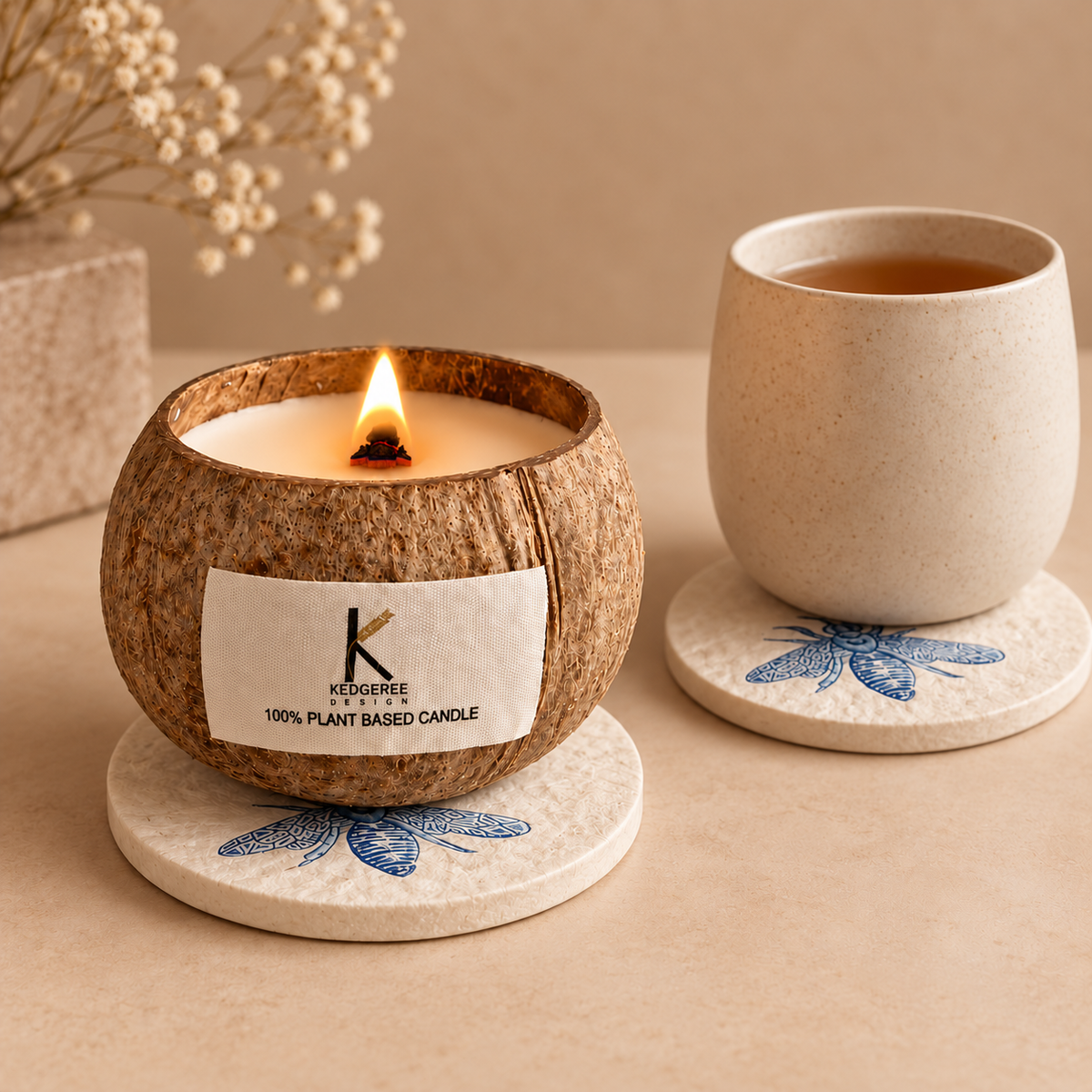 A coconut-shell candle with a lit flame sits on a round coaster beside a matching cup and coaster in a warm beige setting. The candle label reads KEDGEREE CANDLE 100% PLANT BASED CANDLE.