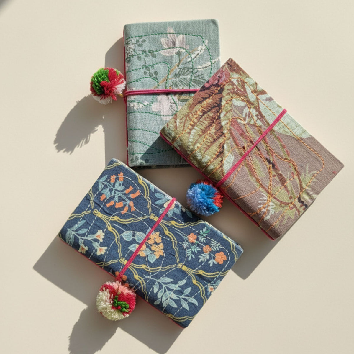 Three decorative fabric-covered notebooks or journals arranged diagonally on a warm beige background. Each journal has a different embroidered floral and botanical pattern in muted tones, tied with a pink elastic band, and decorated with a small pom-pom charm.