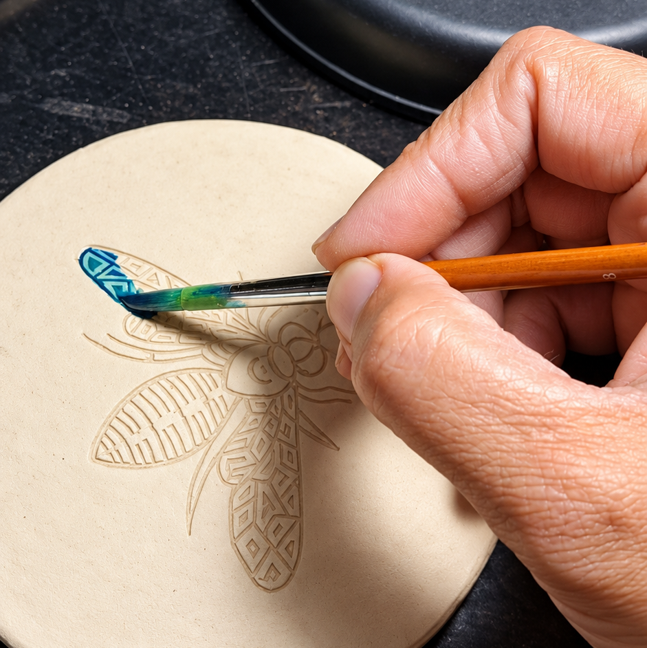 A close-up of a hand holding a small paintbrush and painting a bee design onto a round beige paper mache coaster. A black lamp base is visible in the background on a dark surface.