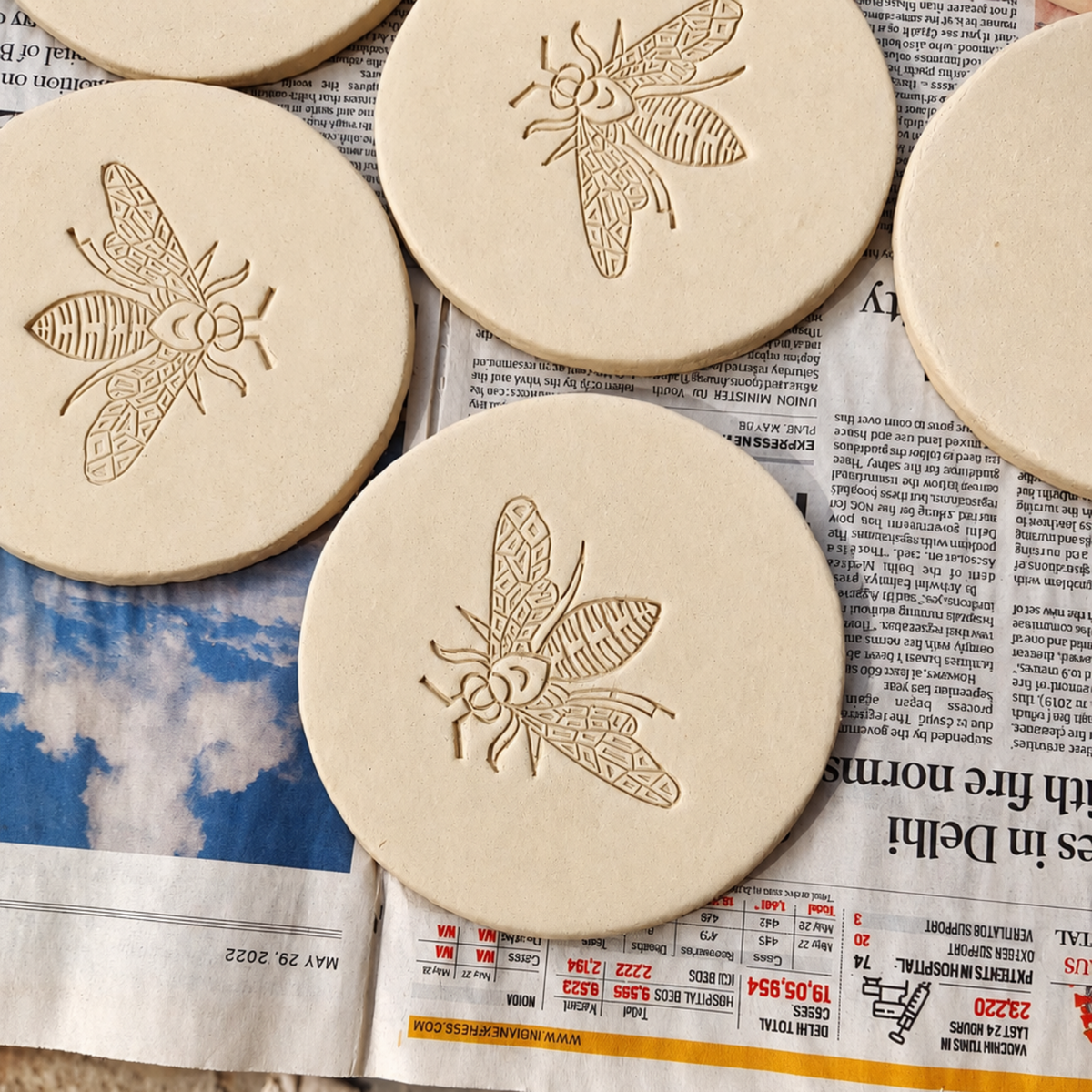 Several round natural kedgeree coasters with engraved bee designs are arranged on top of a newspaper outdoors.