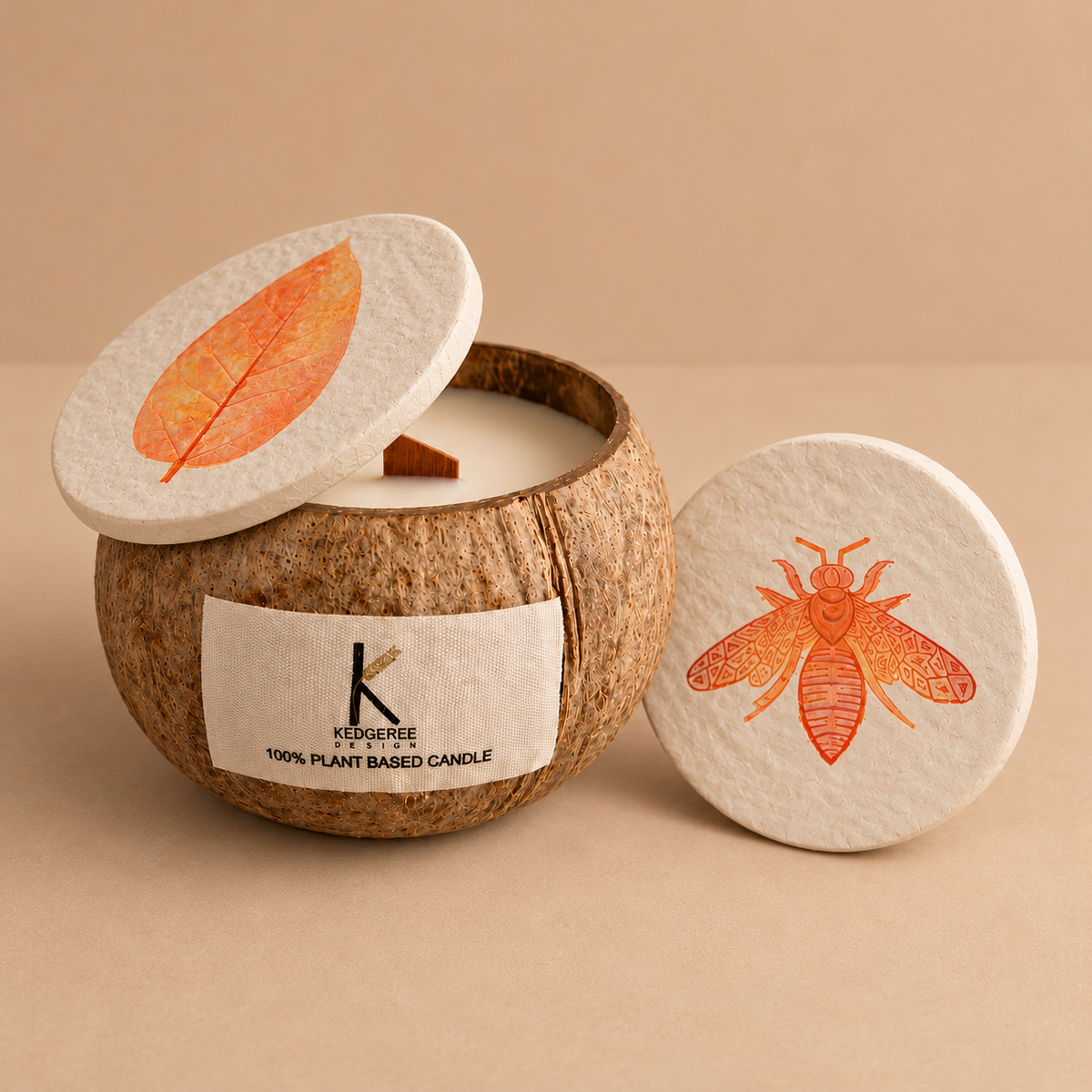 A coconut shell candle jar with a white wax fill, a white label reading Kedgeree CANDLE, and a matching white lid printed with an orange leaf design, shown on a beige background with a second lid featuring an orange bee design beside it.
