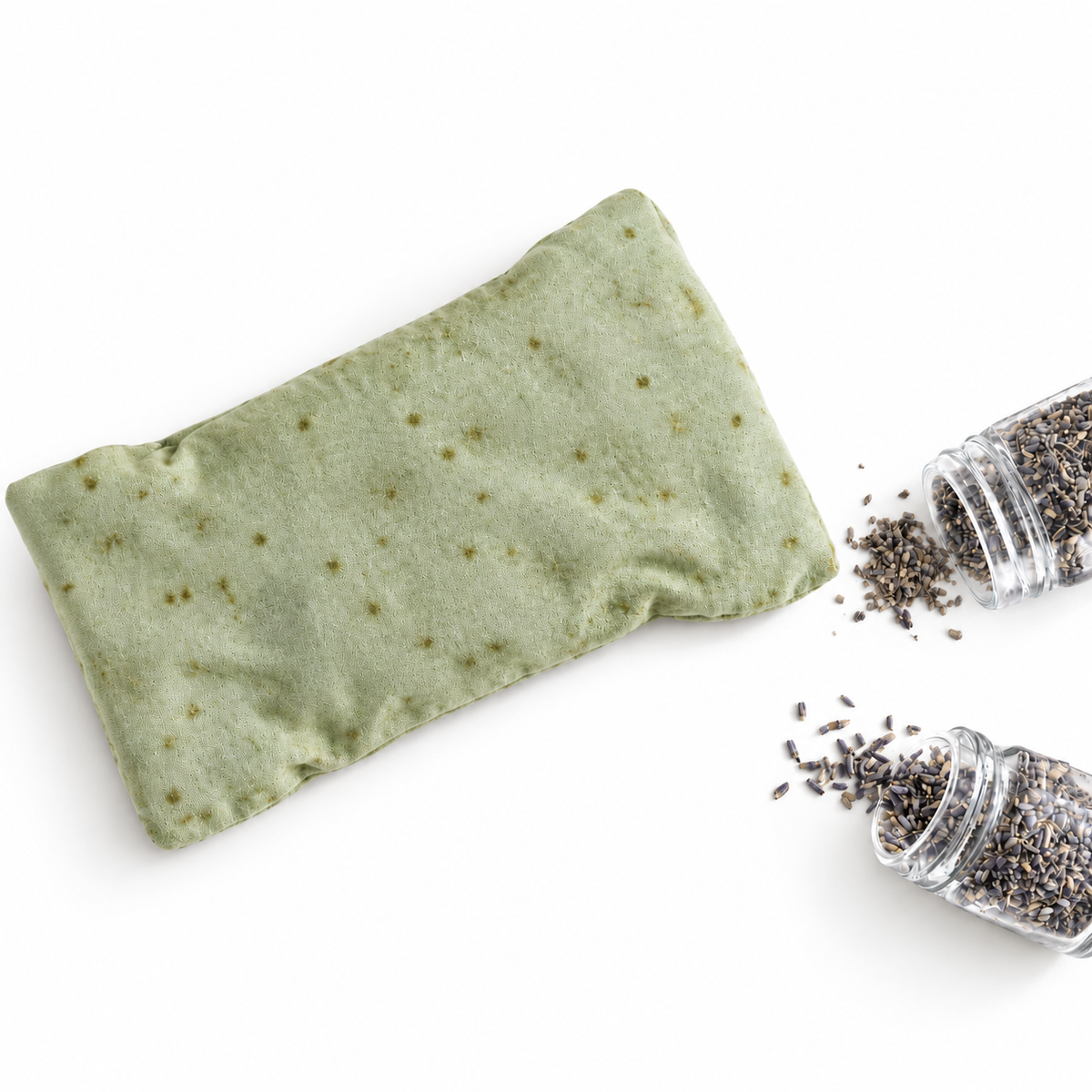 A light sage green fabric eye mask pillow with small embroidered star details, shown next to two clear jars of dried lavender buds on a white background.