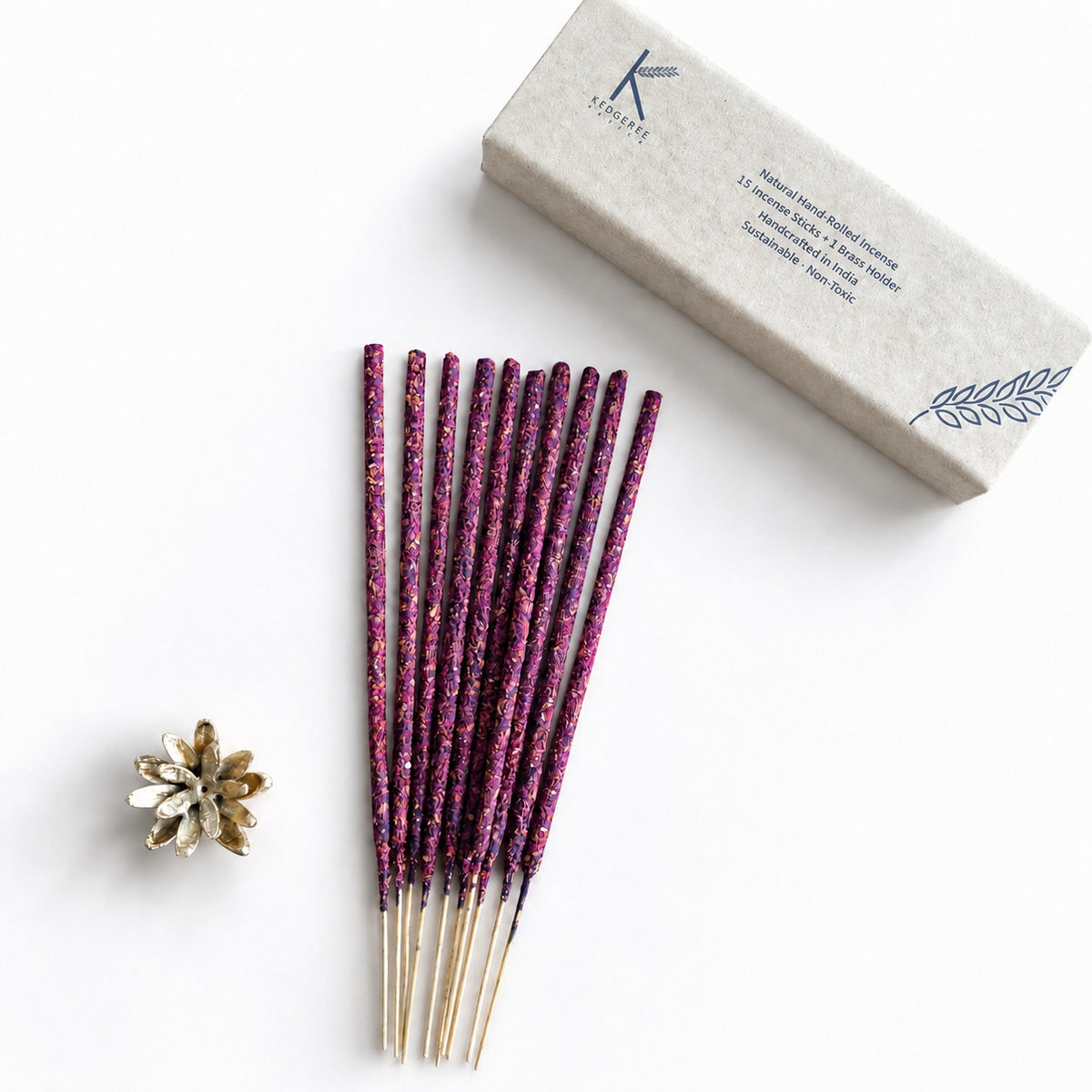 A set of rose handrolled natural  incense sticks arranged next to their textured beige packaging box and a small brass gold incense holder on a clean white background.