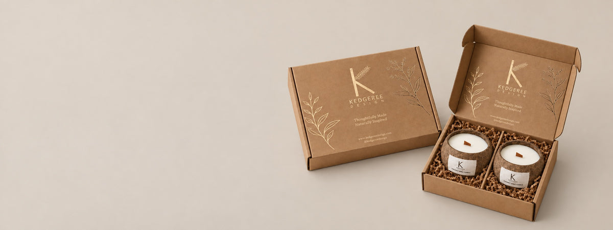 Two coconut shell natural hand poured vegan candles wrapped elegantly in a kraft paper Kedgeree Design gift box with an additional closed matching box beside it on a warm beige background. 
